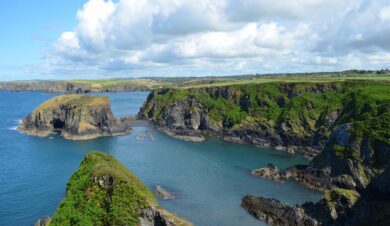 A landscape image of the Pembrokeshire coast with cliffs, interesting geology, green grass and the sea is blue. Also the sky looks lovely with big fluffy white clouds. This scene in near to Old School Bunkhouse