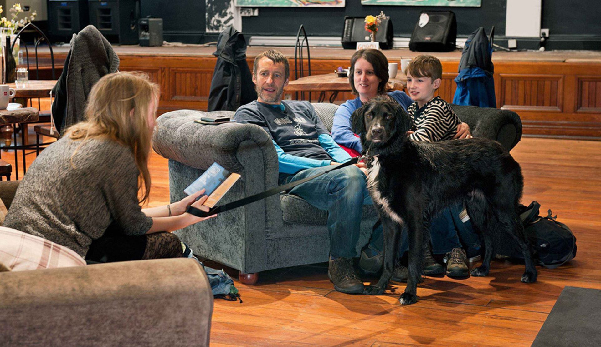 A family with a dog relax on a sofa in the communal area of Cell B hostel. They are chatting to a woman sitting on a nearby armchair. 