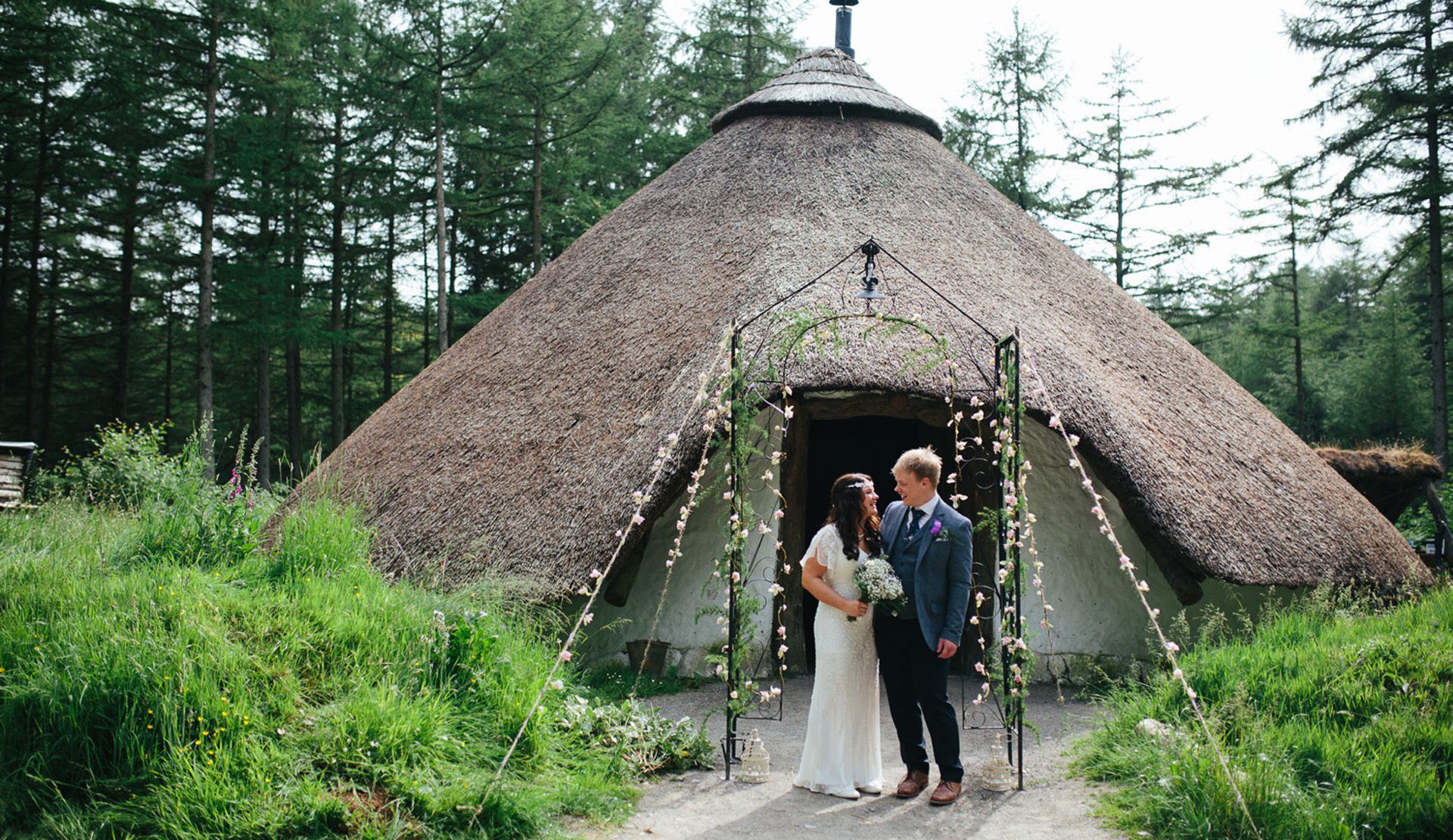 A young couple on their marriage day stood in front of a thatched glamping house