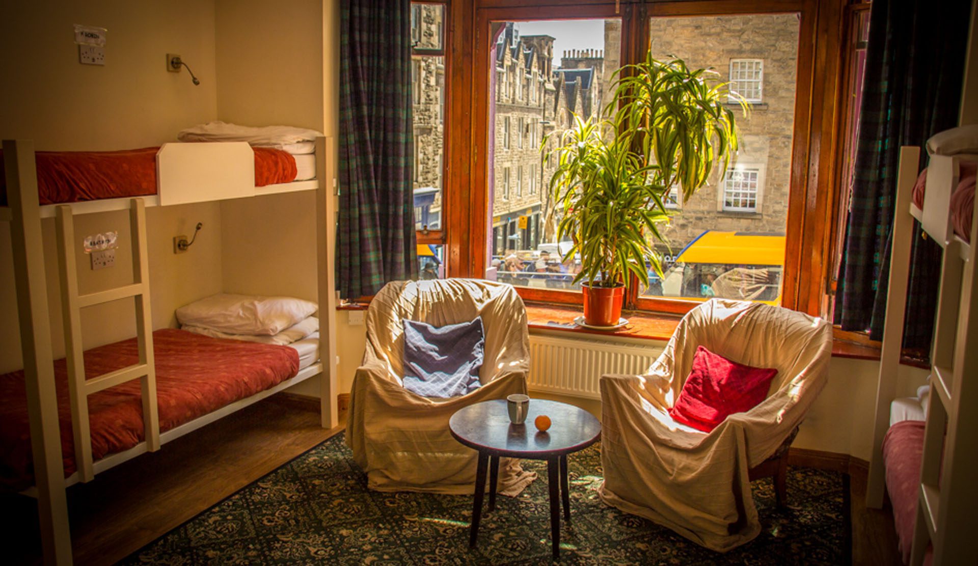 This image depects the sleeping arrangements at Royal Mile backpackers in Edinburgh. There are cosy looking bunk beds and a plant in the big window and chairs with cushions to sit on 
