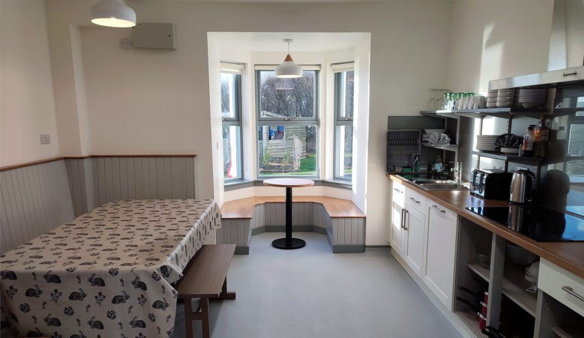 Bright kitchen with large refectory table and small round table in bay window overlooking the graden at Grinneabhat Hostel on the Isle of Lewis