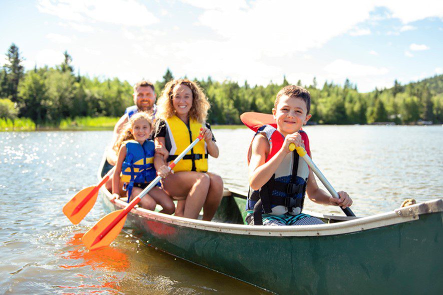 Children ana adults enjoying paddleing a canoe on a large body of water. They are life jackets on and they are all smiling